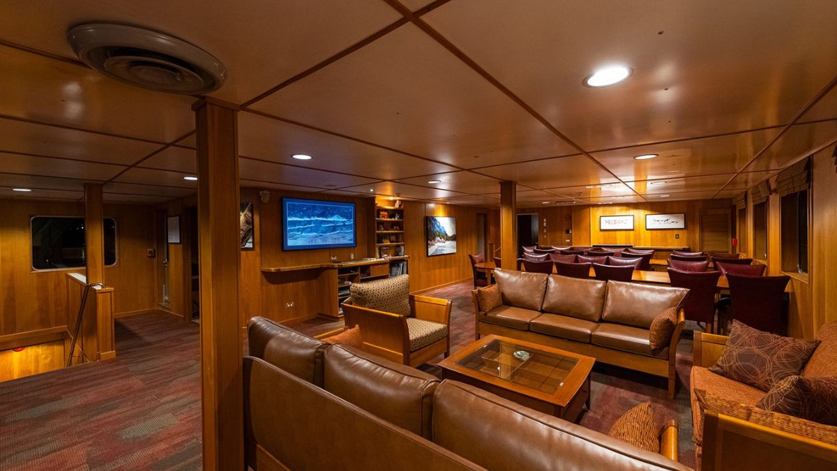 Interior of a cozy lounge area with wooden paneling, featuring comfortable leather sofas, a coffee table, a large screen on the wall, and shelves filled with books and decor. The space is well-lit and designed for relaxation and conversation.