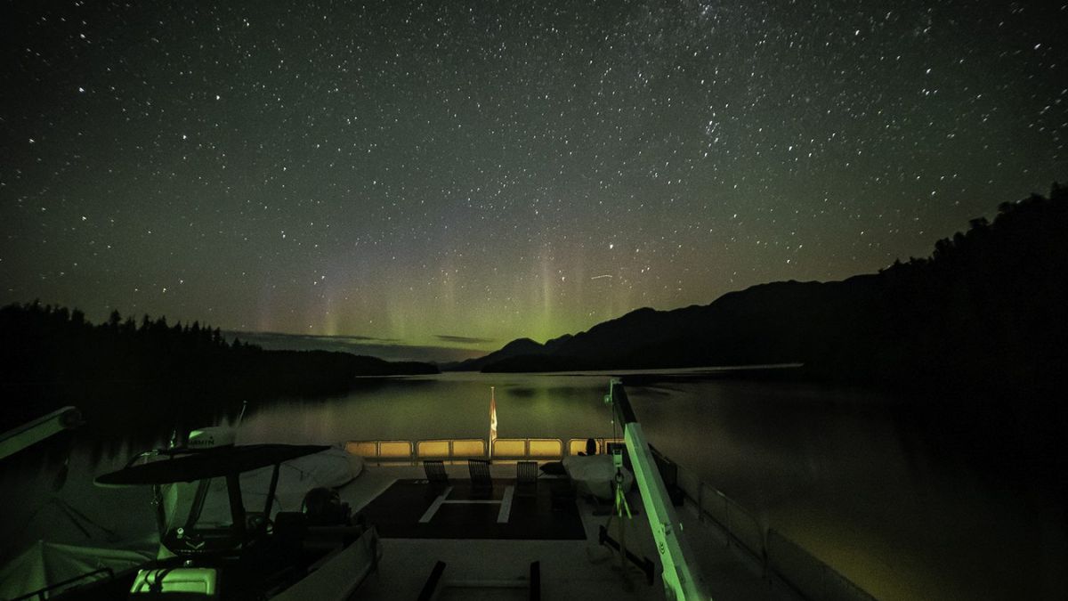 A tranquil night scene over a still lake, illuminated by stars and a faint green aurora in the sky. The silhouette of mountains is visible in the background, and a boat's deck is seen in the foreground.