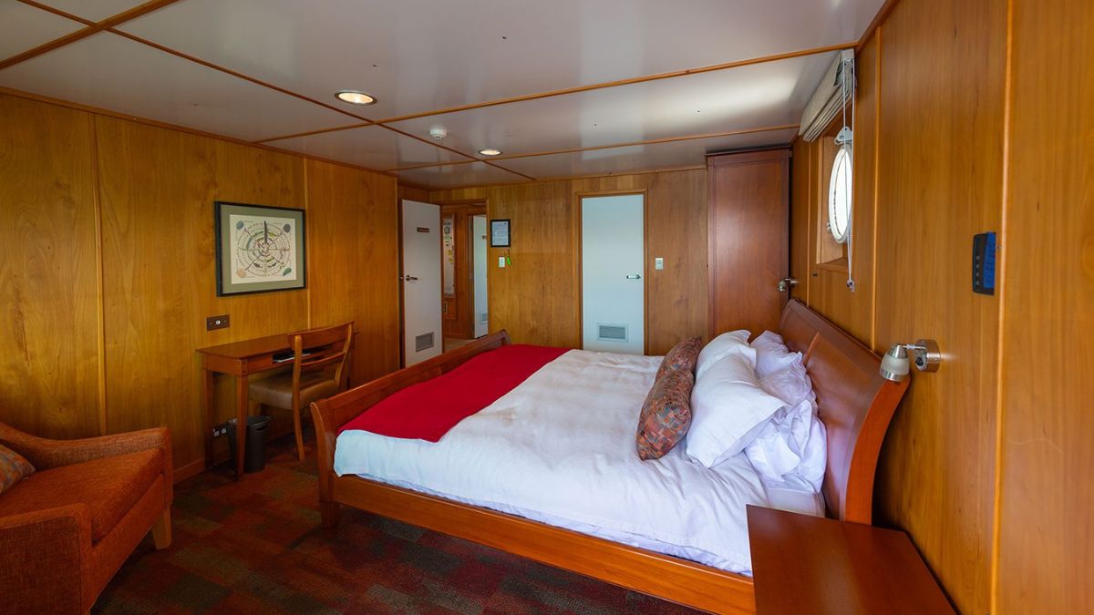 A cozy cabin featuring wooden paneling, a double bed with white linens and a red throw, a small desk and chair, and a round window. A door leads to a bathroom, and another door is opposite the bed.