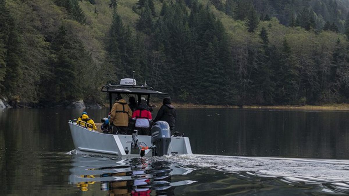A small boat with several people onboard navigates a calm waterway surrounded by lush green forests. The boat leaves a slight wake as it moves through the tranquil scene.
