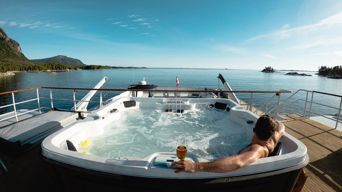 A person relaxes in a hot tub on a yacht, overlooking a serene body of water with islands in the distance. The scene features clear blue skies and lush green forests along the shoreline. A drink is held in one hand, enhancing the tranquil atmosphere.