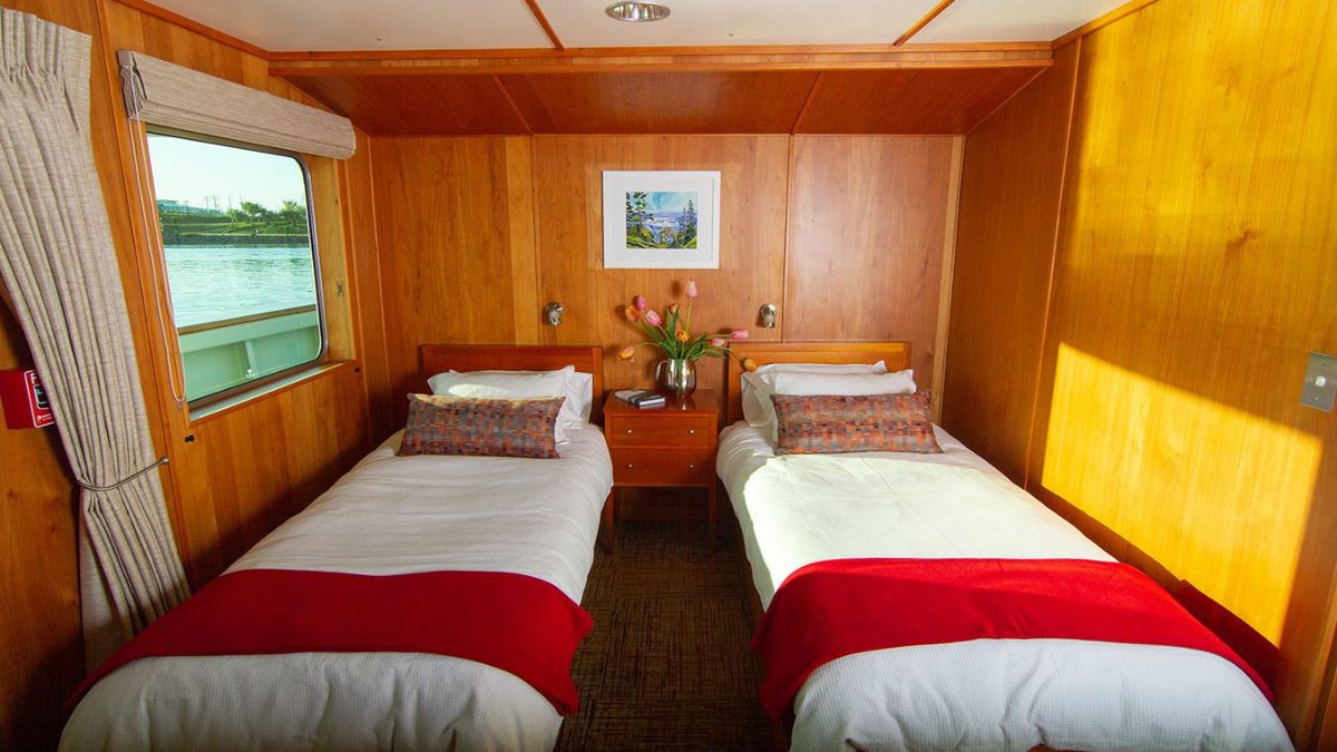 Cozy cabin interior featuring two twin beds with white linens and red throws. A wooden wall creates a warm ambiance, complemented by a small side table with flowers and a framed landscape artwork. Natural light filters through a window.