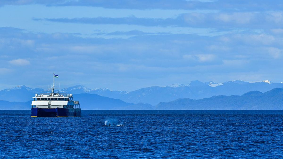 A ferry boat sails on a calm blue ocean, with distant mountain ranges and a clear sky in the background.