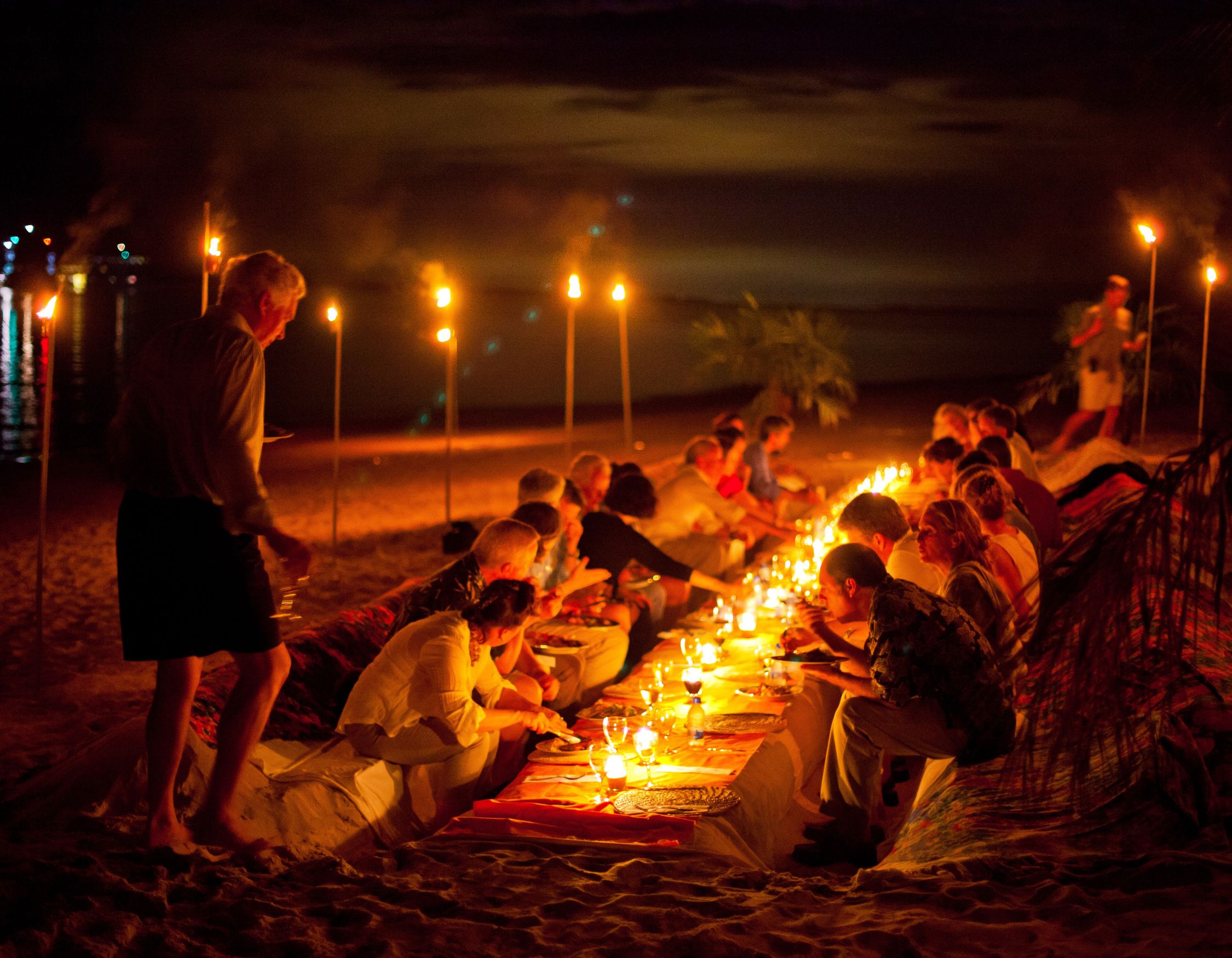 A large group of people dining at a long candlelit table on a beach at night under tiki torches.