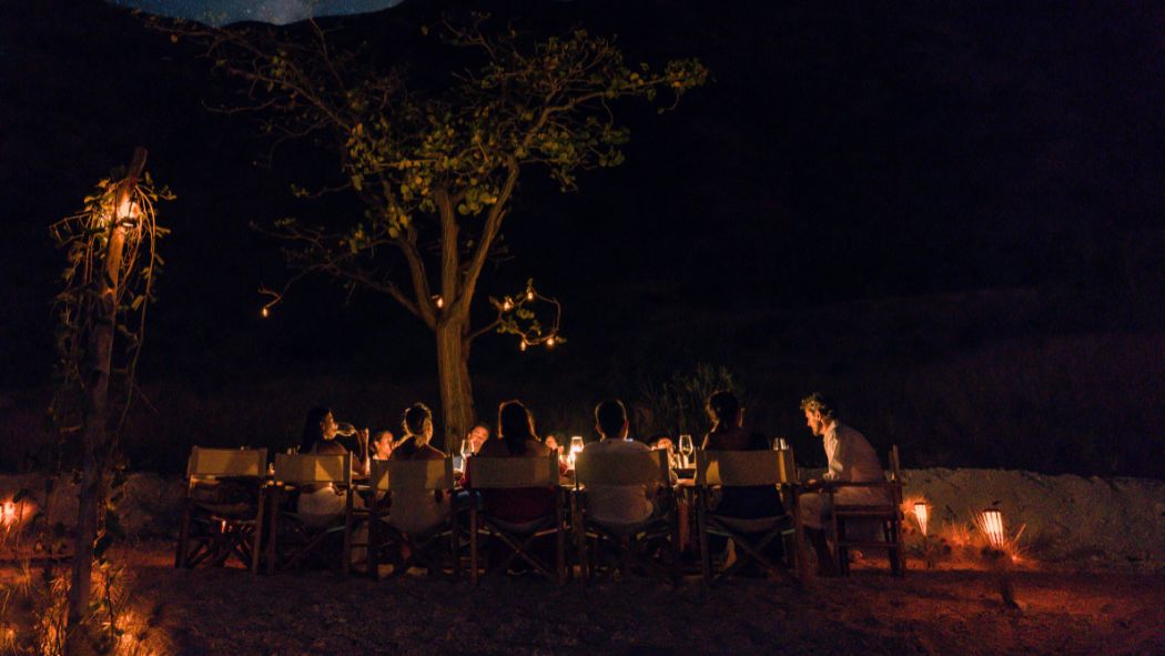 A group of people gathered around a table outdoors at night, illuminated by lanterns and string lights. A large tree is positioned in the background, creating a cozy atmosphere.