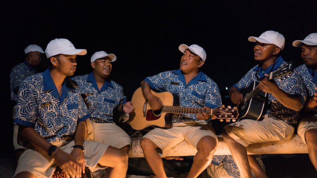A group of men in matching blue patterned shirts and white hats sings and plays guitars around a campfire at night. Their joyful expressions reflect a lively atmosphere.