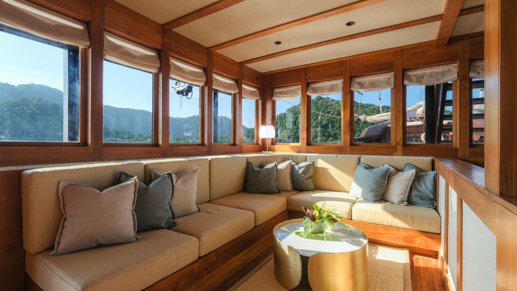 A cozy, well-lit lounge area features a U-shaped beige sofa adorned with decorative pillows, surrounded by large windows that offer scenic views of lush green mountains. A small round coffee table with a vase of flowers sits in the center.