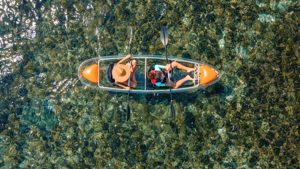 Two people in a kayak paddling over clear, shallow water filled with visible rocks and aquatic plants. One person wears a wide-brimmed hat and the other is in a red shirt. Sunlight reflects off the waters surface.