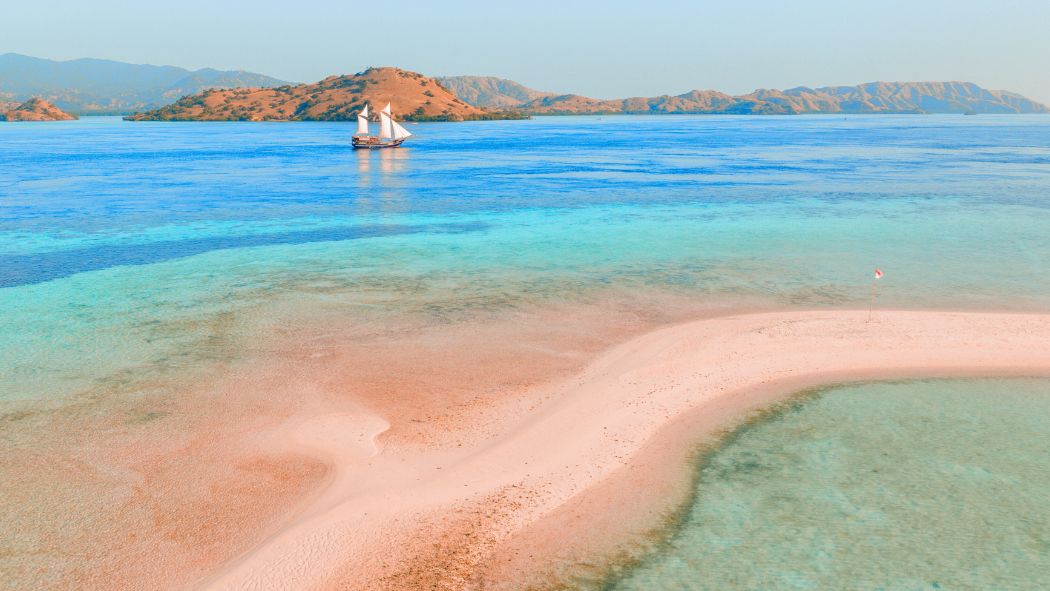 A serene coastal landscape featuring a sandy peninsula extending into clear turquoise waters, with green islands in the background and a sailboat gently gliding across the sea.