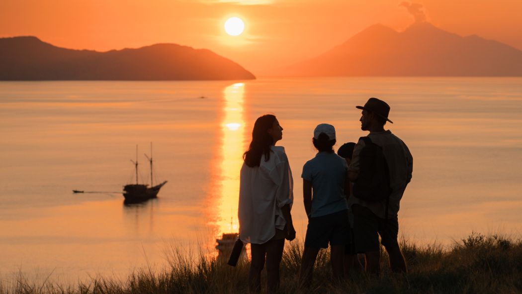 A group of four people stands on a hilltop, silhouetted against a vibrant sunset over calm waters. The sun is low on the horizon, casting golden reflections on the water, while distant mountains create a scenic backdrop. A boat is visible in the foreground.