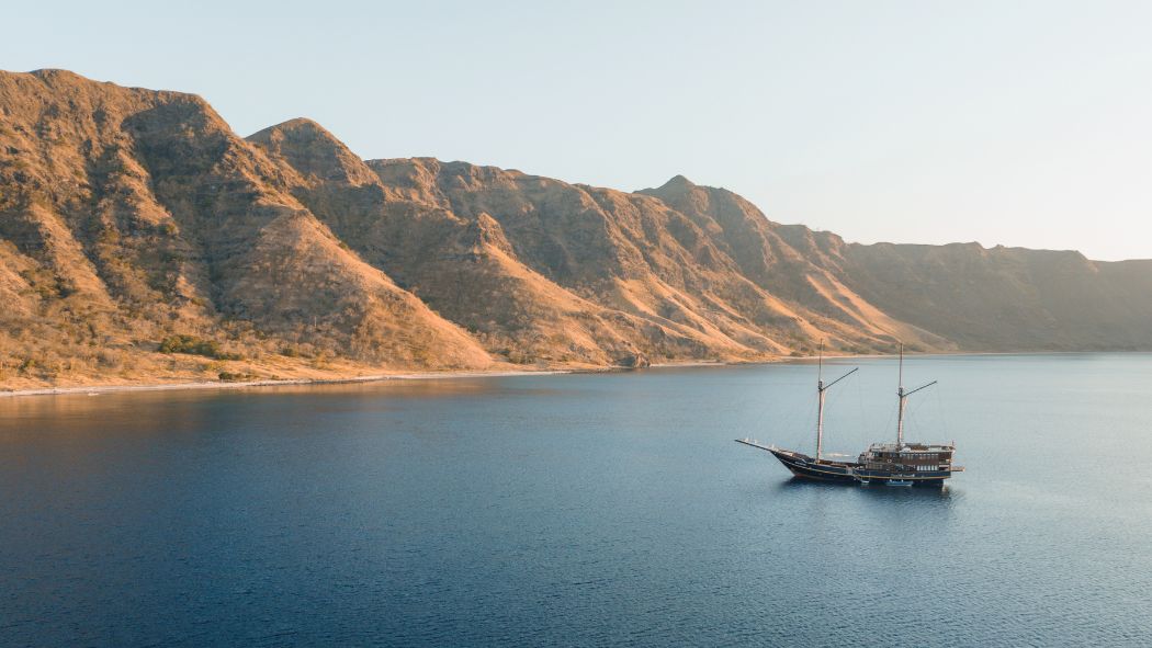 A traditional sailing ship anchored in calm blue water, with rugged, sunlit mountains rising in the background.