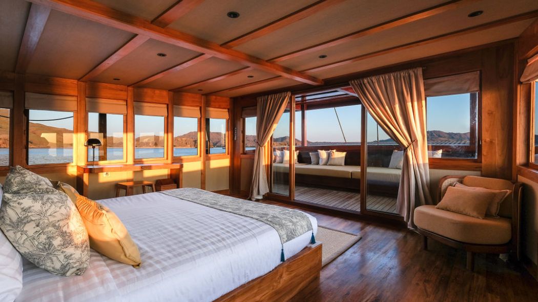 A beautifully designed bedroom with wooden beams and large windows offering a view of the water. The room features a comfortable bed with decorative pillows, a cozy armchair, and light-colored curtains. Sunlight fills the space, highlighting the warm tones of the wood and furnishings.