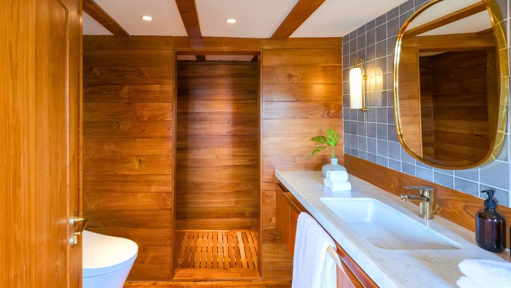 A modern bathroom featuring wooden walls and a shower area with a wooden slatted floor. The sink area has a sleek countertop with a round mirror above, complemented by a gold light fixture. A small plant adds a touch of greenery, and towels are neatly arranged nearby.