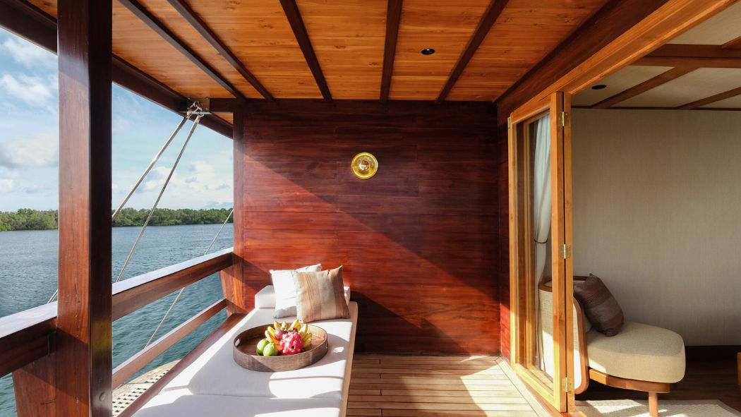A cozy, wooden terrace overlooking water, featuring a lounge chair with cushions and a bowl of fruit on a table. Sunlight streams through a nearby glass door leading into a stylish interior.