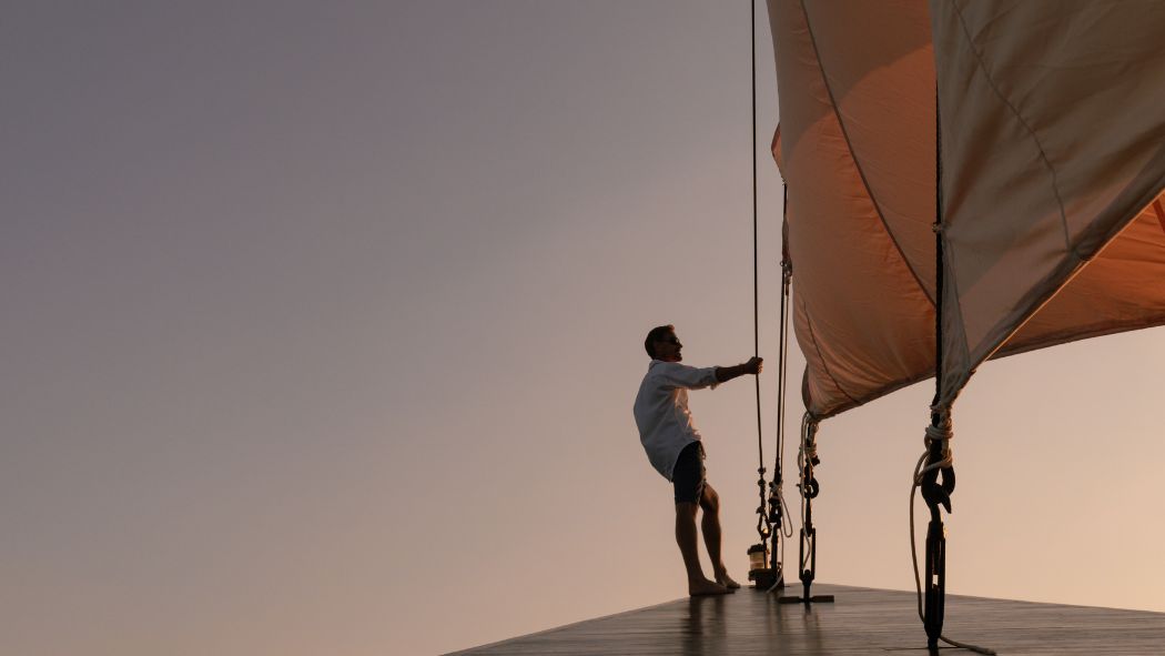 A person stands on the bow of a sailboat, pulling on the sail rigging against a backdrop of a sunset sky. The sail is billowing in the wind, and the scene conveys a sense of adventure and tranquility.