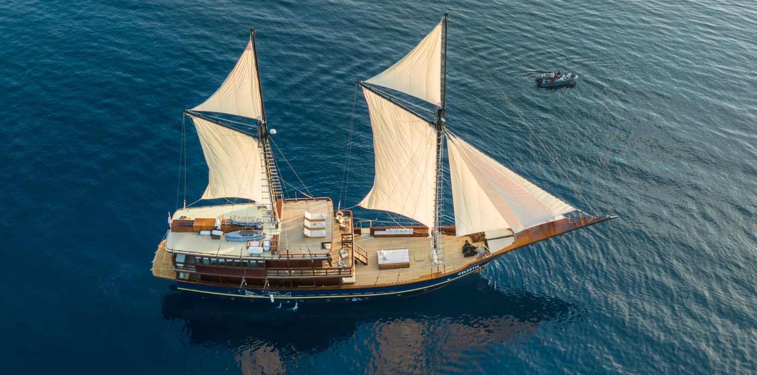 A large sailing ship with two prominent sails glides through calm blue waters, surrounded by a smaller boat nearby. The ship features a wooden deck and a variety of seating areas.