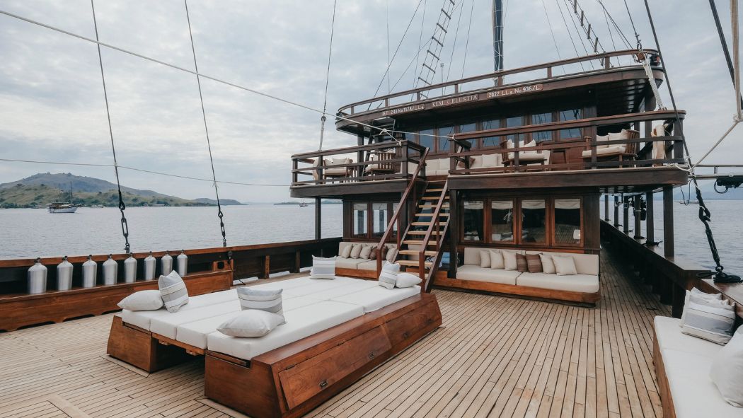 A spacious wooden deck of a yacht features lounge seating and a staircase leading to an upper deck. The serene sea stretches into the distance with a faint outline of an island.