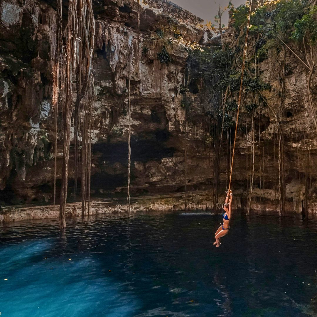 A person swings from a rope over a deep blue cenote surrounded by rocky cliffs and lush greenery. Sunlight filters through the trees above, illuminating the tranquil water below.