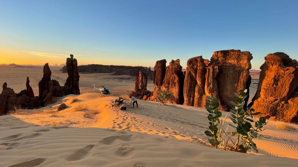 A scenic desert landscape at sunset, featuring towering rock formations and gentle sand dunes. A helicopter is parked nearby, with people gathered around on the sandy terrain. Lush green plants contrast with the warm tones of the sand and rocks.