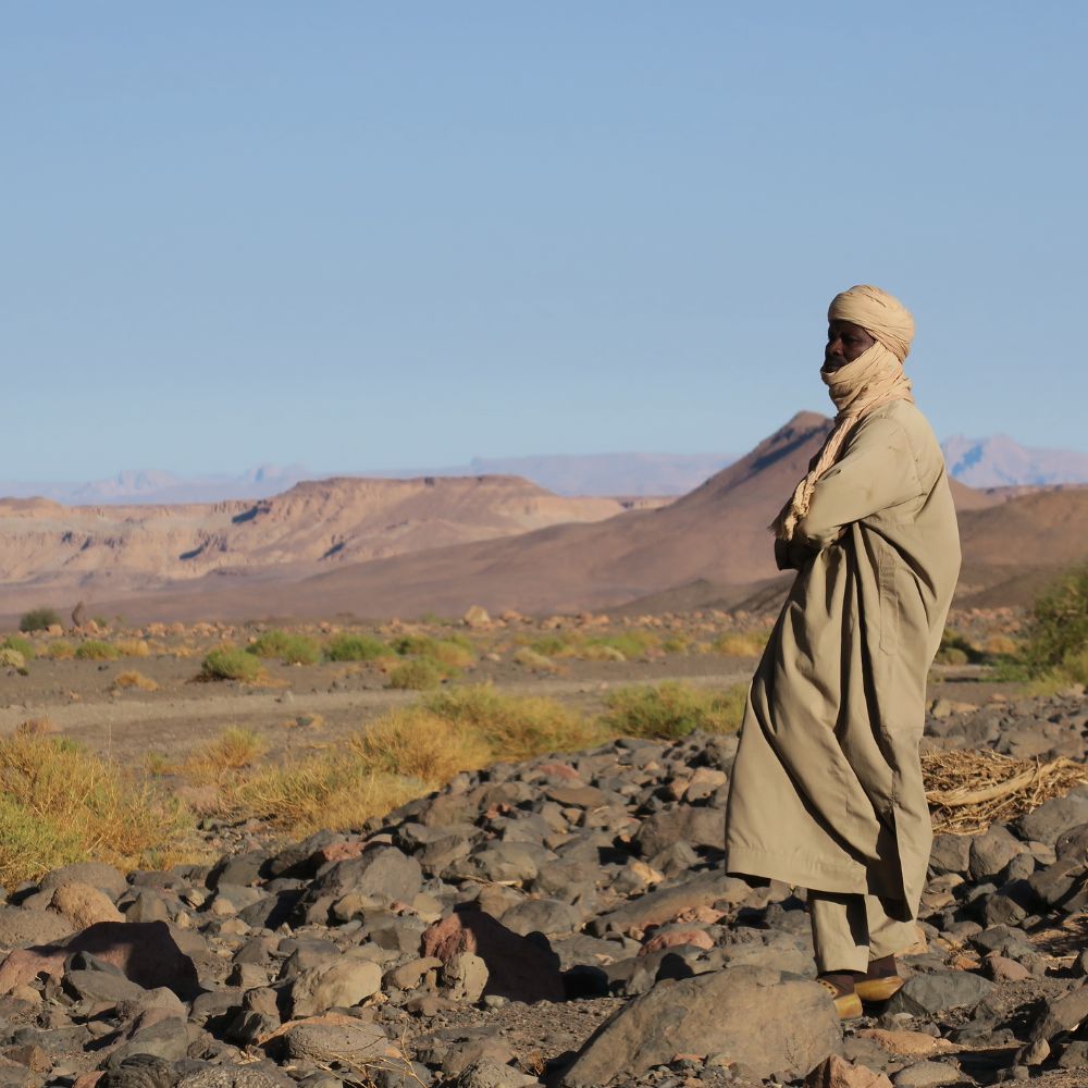 A person wearing traditional clothing stands on rocky terrain with a vast landscape of mountains and sparse vegetation in the background. The sky is clear and blue.