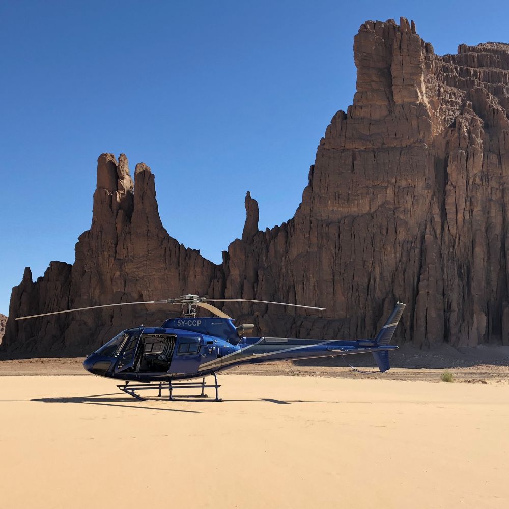 A blue helicopter is parked on a sandy desert landscape, with tall, rugged rock formations in the background under a clear blue sky.