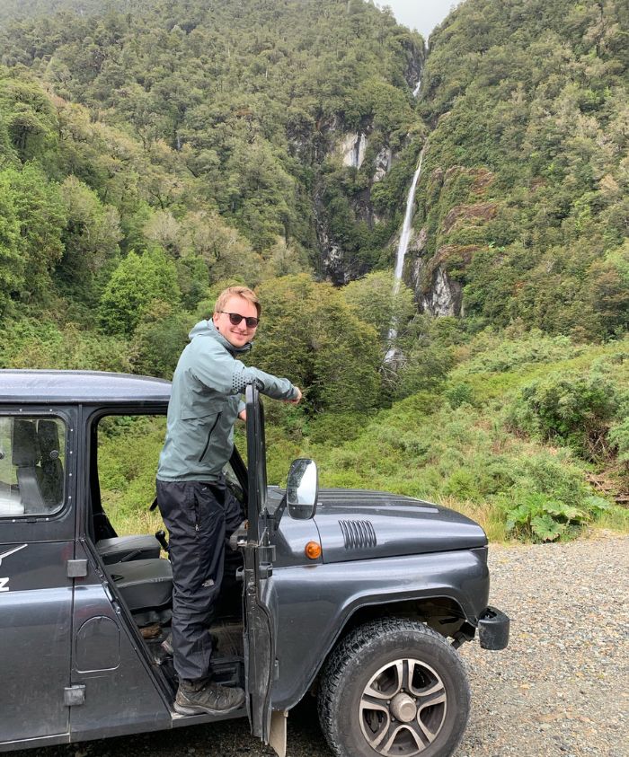 A person stands by a parked off-road vehicle in a lush green forest, smiling at the camera. In the background, a tall waterfall cascades down a rocky cliff.
