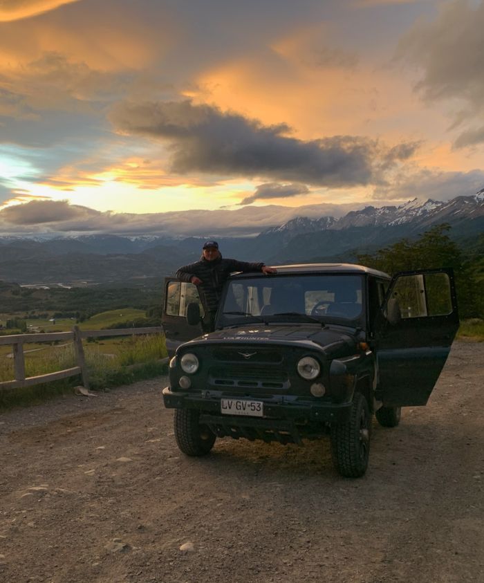 A person leans on a black off-road vehicle parked on a gravel road, with dramatic clouds and a colourful sunset in the background. Snow-capped mountains and lush green valleys are visible in the distance.