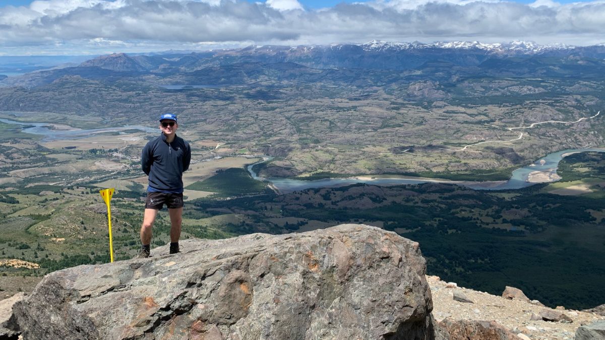 A person stands on a rocky outcrop at a mountain summit, wearing shorts and a hat. The vast landscape below features lush valleys, winding rivers, and distant snow-capped mountains under a partly cloudy sky.