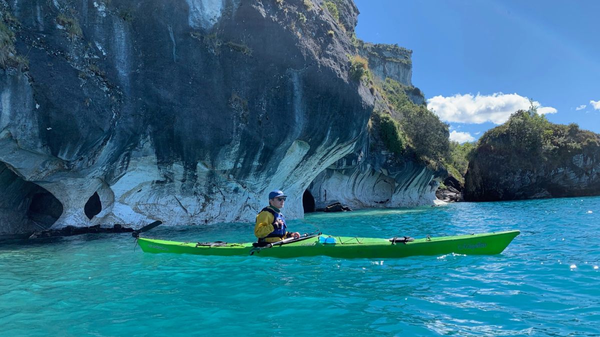 A person in a yellow jacket kayaking in turquoise water near striking rock formations with caves in the background. The scene is bright and sunny with a clear blue sky.