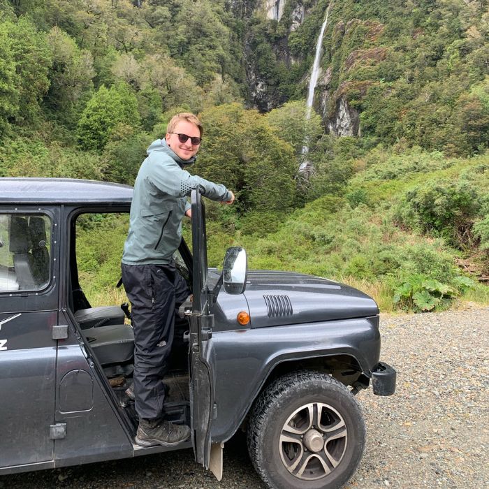 A person wearing sunglasses and a green jacket leans out of the driver's side of a rugged car, with a waterfall and lush greenery in the background.