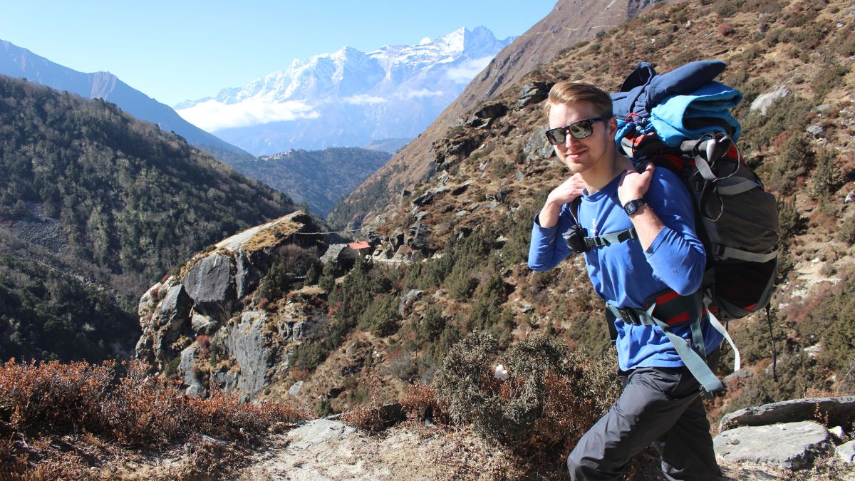 A young man wearing sunglasses carries a large backpack while hiking along a rocky trail, flanked by steep hills and snow-capped mountains in the background.
