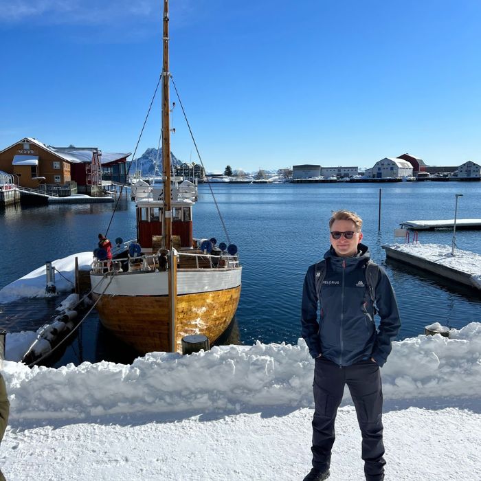 A young man stands on a snowy dock beside a wooden boat in a harbor. The background features calm water and buildings under a clear blue sky.