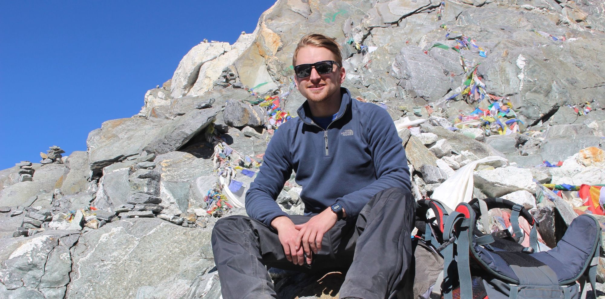 A young man wearing sunglasses sits on rocky terrain, surrounded by colorful prayer flags and stones. He is dressed in a blue fleece jacket and gray pants, looking relaxed against a backdrop of clear blue sky and rugged mountain rocks.