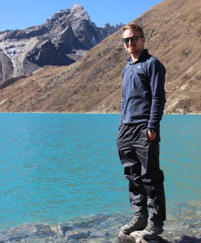 A person stands on a rocky shore beside a turquoise lake, with rugged mountains rising in the background. They are wearing a navy fleece top and dark trousers, along with sunglasses. Bright sunlight illuminates the scene.
