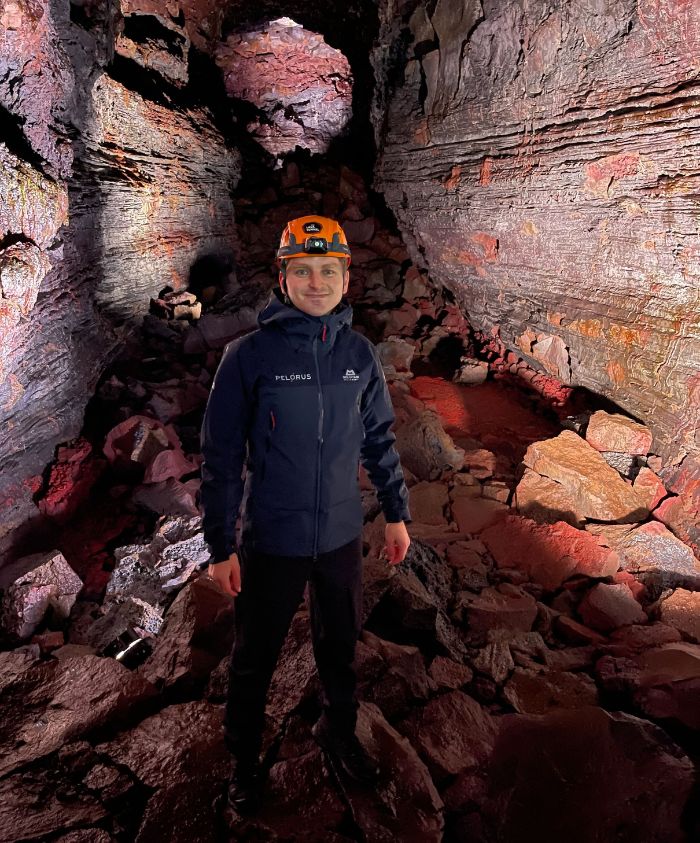 A person wearing a helmet and a jacket stands among rocky terrain inside a dark, volcanic cave. The walls display colorful layers of rock, illuminated by soft red and purple lighting.
