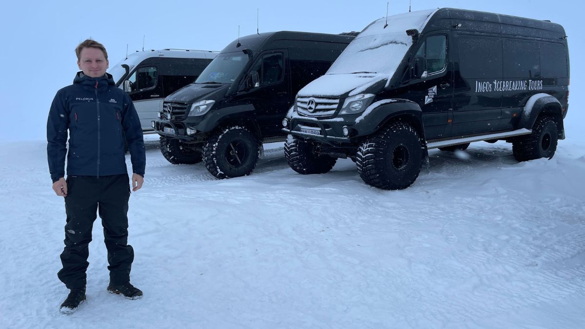A person stands in front of three rugged black vans parked on a snowy landscape. The vans are modified for winter conditions, featuring large tires and snow-covered exteriors. The setting appears cold and wintry.