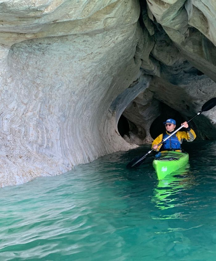 A person in a green kayak paddles through a narrow, rocky cave with smooth, swirling walls. The water is turquoise and clear, reflecting the cave's intricate textures.