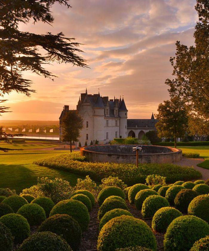 A picturesque landscape featuring a chateau surrounded by manicured gardens and neatly trimmed hedges, with trees and a golden sunset illuminating the scene.