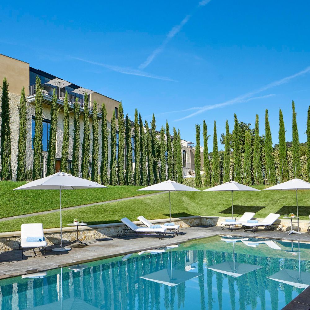 A luxurious swimming pool surrounded by tall, green cypress trees and lounge chairs with umbrellas. A modern building is visible in the background under a clear blue sky.