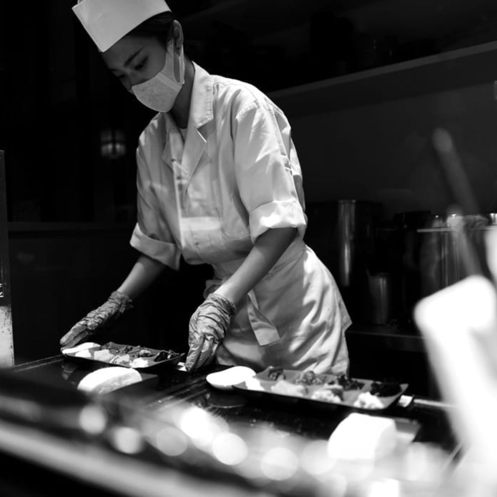 A chef in a white uniform and mask carefully arranges sushi on two plates in a dimly lit kitchen.
