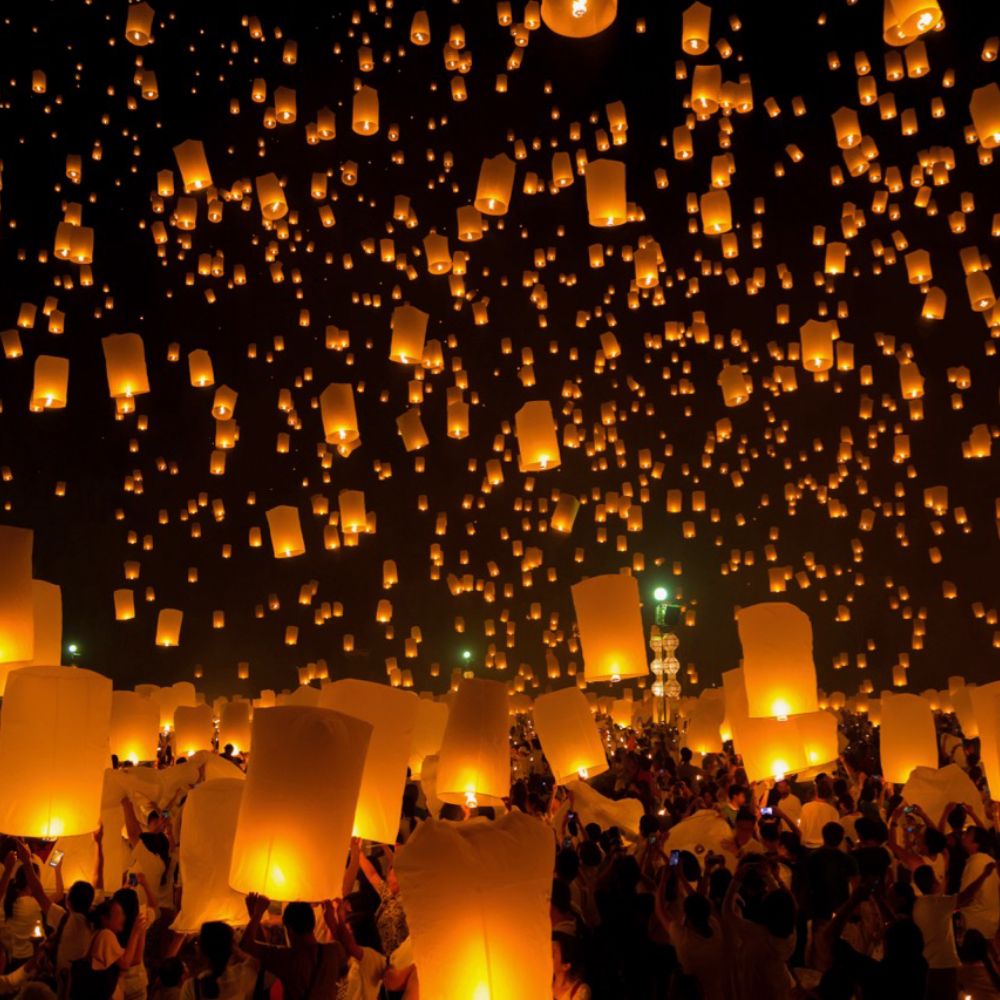 Hundreds of glowing orange sky lanterns floating in the dark night sky above a crowded festival.