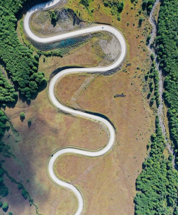 Aerial view of a winding road cutting through lush green trees and golden grasslands. The road forms a series of curves, leading into the distance.
