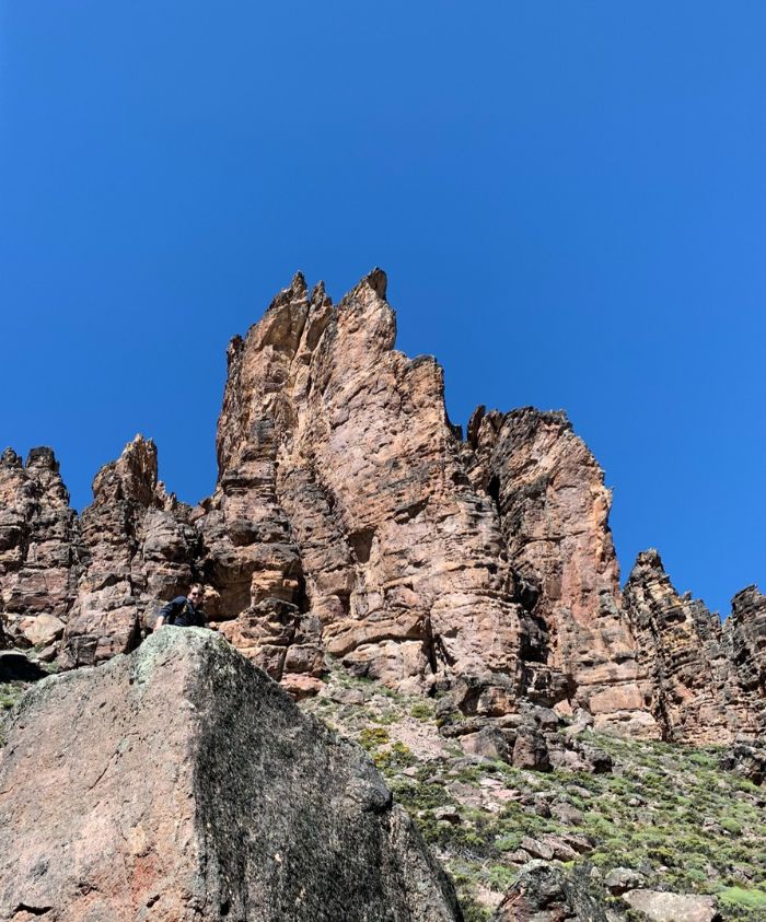 A towering rock formation rises against a clear blue sky, with rugged cliffs and green vegetation at the base. A small figure stands on a rock, highlighting the scale of the landscape.