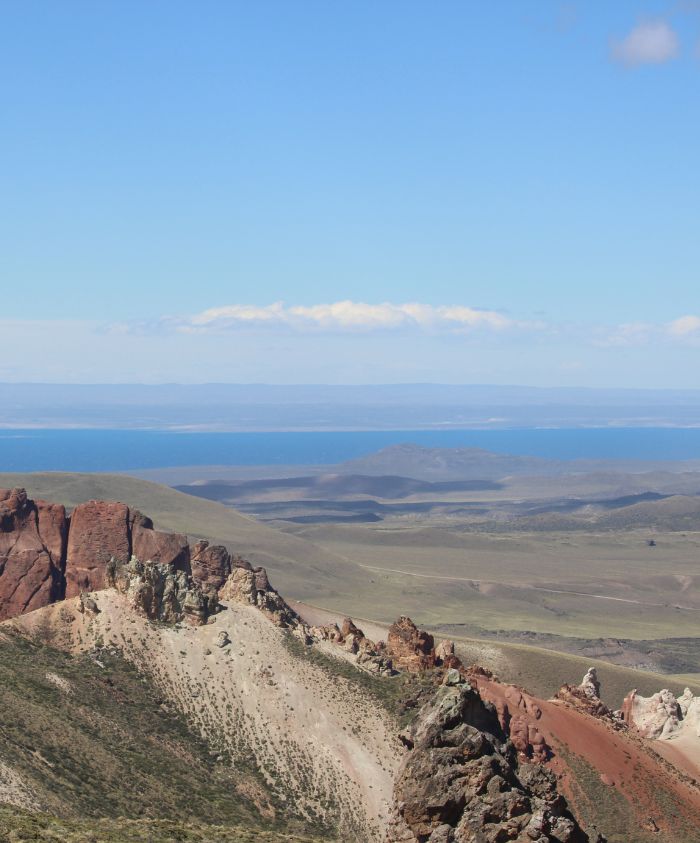 A panoramic view of rugged mountain terrain with reddish rock formations in the foreground, transitioning into green hills, and a distant tranquil blue sea under a clear sky.