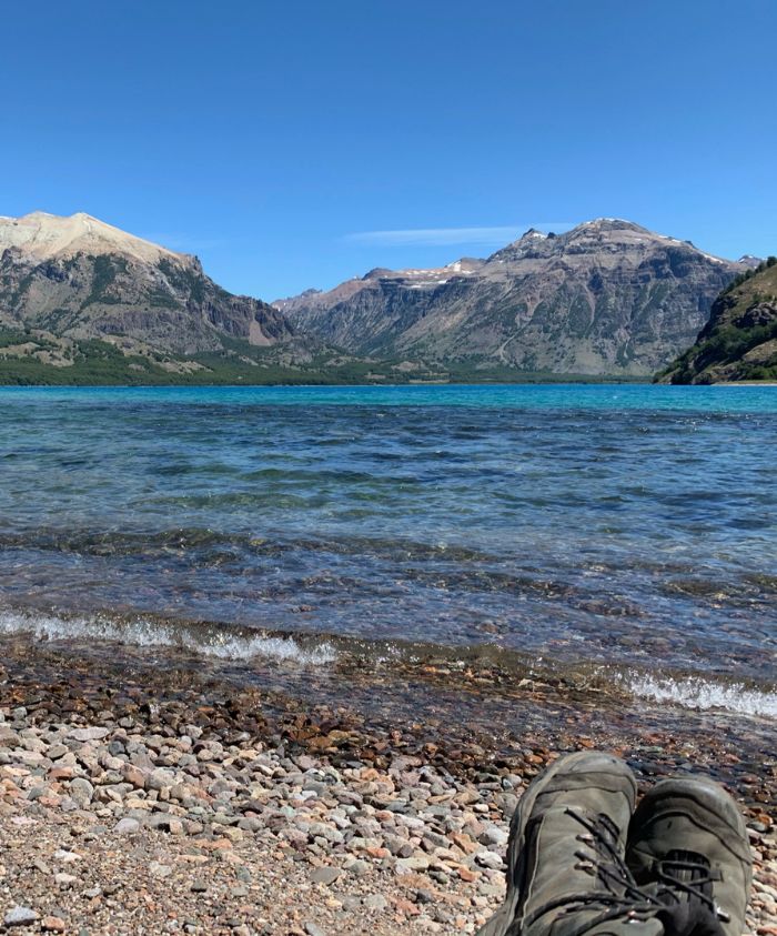 A pair of hiking boots resting on a rocky shore with clear blue water in the foreground. Majestic mountains rise in the background under a bright blue sky.