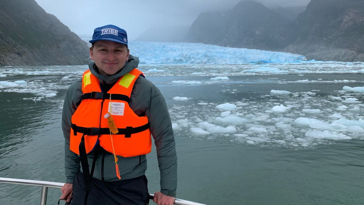 A man in a bright orange life vest smiles while standing on a boat, with a glacier and large icebergs visible in the background. The scene is set in a misty, mountainous environment.