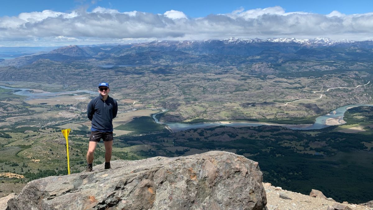 Person standing on a rocky outcrop at a mountain summit, wearing a blue cap and dark clothing. A scenic valley and rivers are visible in the background, surrounded by lush greenery and distant snow-capped peaks.