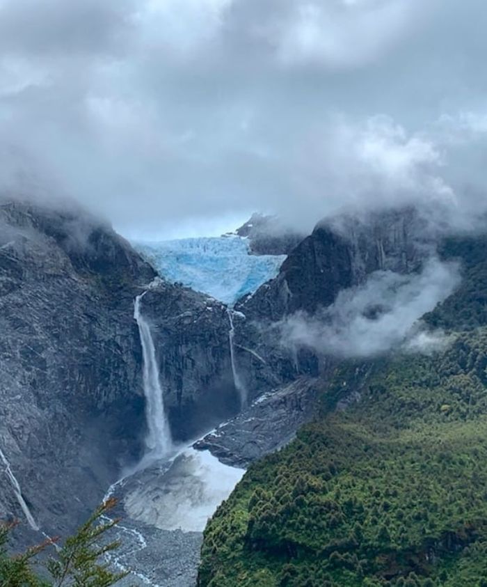 A stunning landscape featuring a glacier in a rocky mountain range, with waterfalls cascading down into a lush green valley. The scene is enveloped in mist and clouds, creating a dramatic atmosphere.