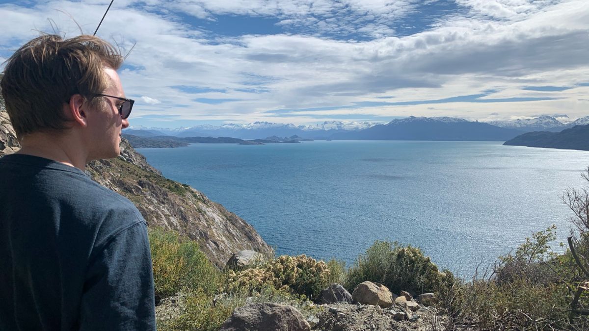 A person wearing sunglasses stands on a rocky coastline, gazing out at a vast blue lake surrounded by mountains under a partly cloudy sky.