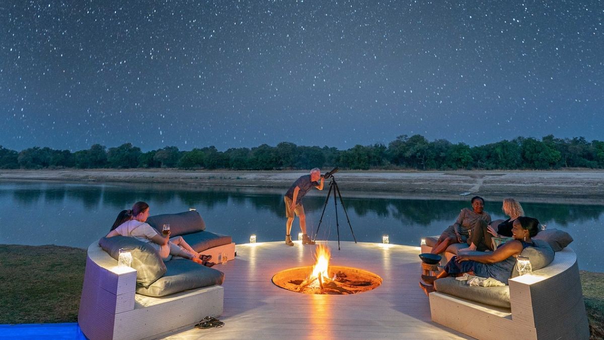 A serene outdoor gathering by a riverbank at night, featuring a campfire surrounded by comfortable seating. A person sets up a camera on a tripod, while others relax and enjoy drinks, under a sky filled with stars.